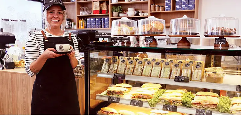 Smiling TCM Market staff in an apron holding a cup of coffee behind a display filled with sandwiches and pastries under cake domes