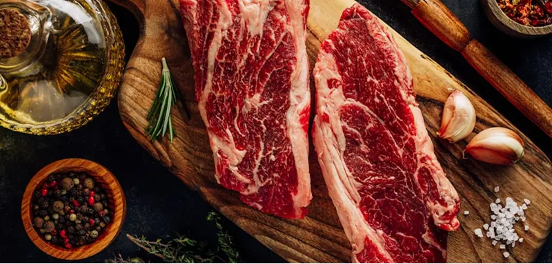 Two raw marbled steaks on a wooden board, surrounded by garlic, herbs, peppercorns, coarse salt, and olive oil.
